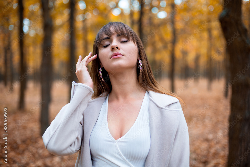 stylish young caucasian female with dark hair in the light coat in autumn park, trees with orange leaves and leaves on the ground stylish young caucasian female with dark hair in the light coat in autumn park, trees with orange leaves and leaves on the ground