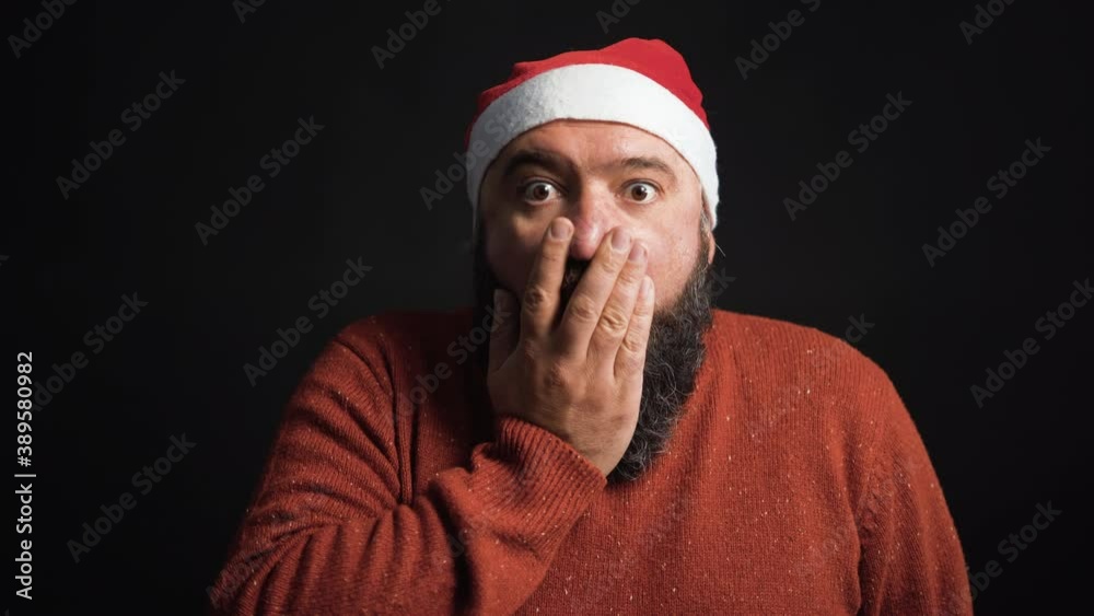 a well-fed bearded man dressed in a Santa Claus hat is surprised and covers his face with his palm Black background. Christmas concept a well-fed bearded man dressed in a Santa Claus hat is surprised and covers his face with his palm Black background. Christmas concept