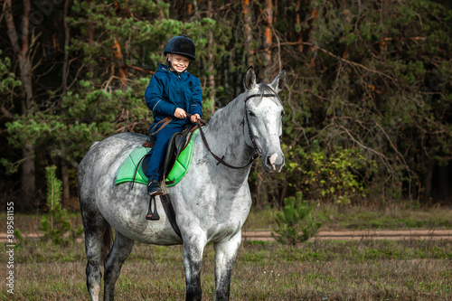Little boy in a jockey cap on a white adult horse on a background of nature. Jockey, hippodrome, horseback riding
