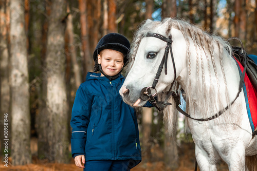 Boy in a jockey cap portrait, stands next to a white pony close-up on the background of nature. Jockey, epodrome, horseback riding.