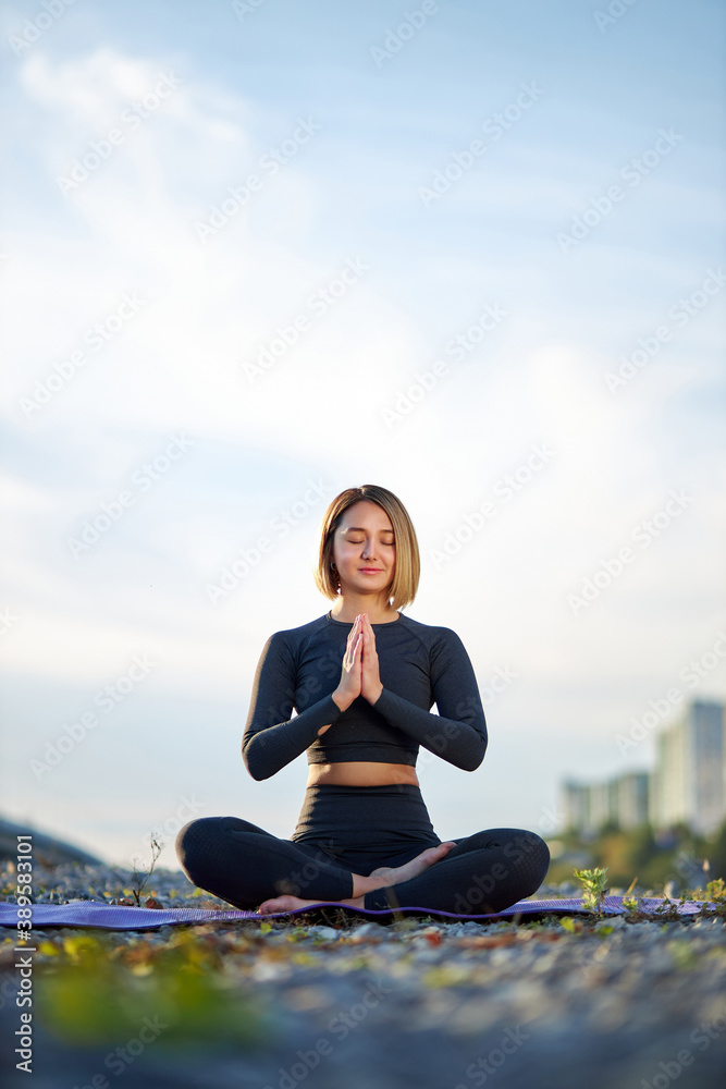 portrait of young calm caucasian female meditating outdoors, beautiful woman in sportswear sit on mat and keep calm, in pose with crossed legs