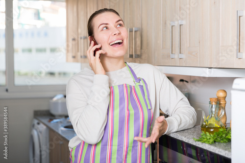 Young woman is talking by phone with girlfriend in the kitchen at the home.