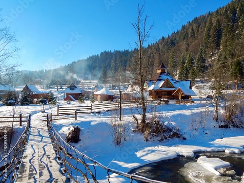 mountain village in winter, snow and wooden houses, suspension bridge across the icy river © Olena