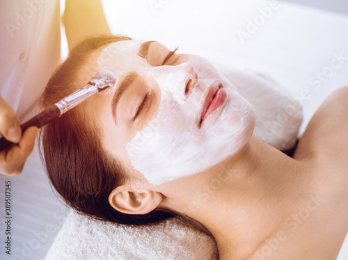 Beautiful brunette woman enjoying applying cosmetic mask with closed eyes in sunny spa center. Relaxing treatment and medicine concept