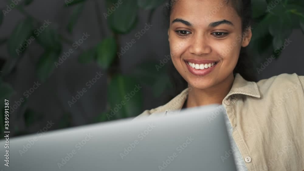 A pleased african american girl is using her laptop computer sitting in the chair indoors in the cafe
