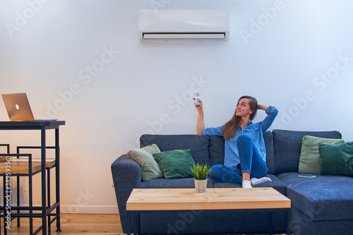 Young happy woman sitting on couch under air conditioner and adjusting comfort temperature with remote control at modern home photo