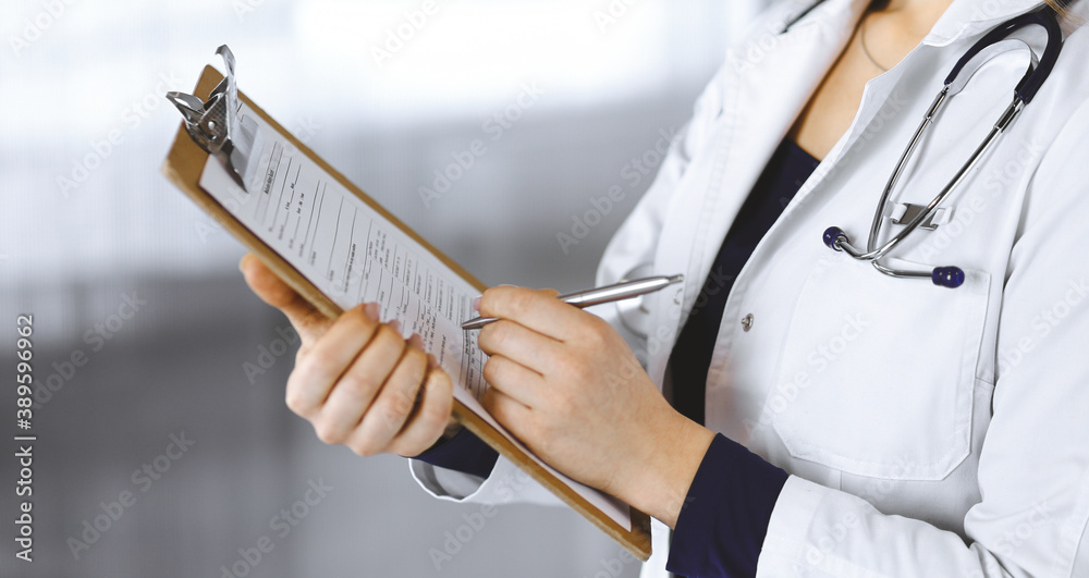 Unknown woman-doctor is writing some notes using a clipboard, while standing in the cabinet in a clinic. Female physician with a stethoscope, close-up. Perfect medical service in a hospital. Medicine