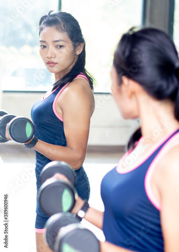 Young woman practicing with dumbbells in front of mirror in gym. Girl strengthens in fitness club.