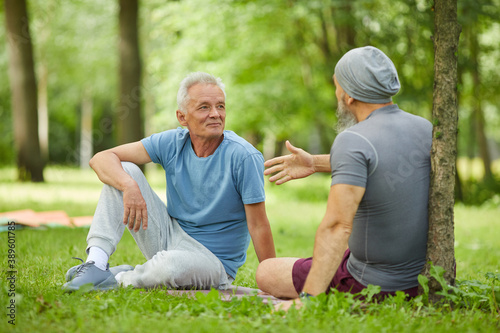 Two sporty senior male friends sitting together on green grass in park talking about something, full body shot