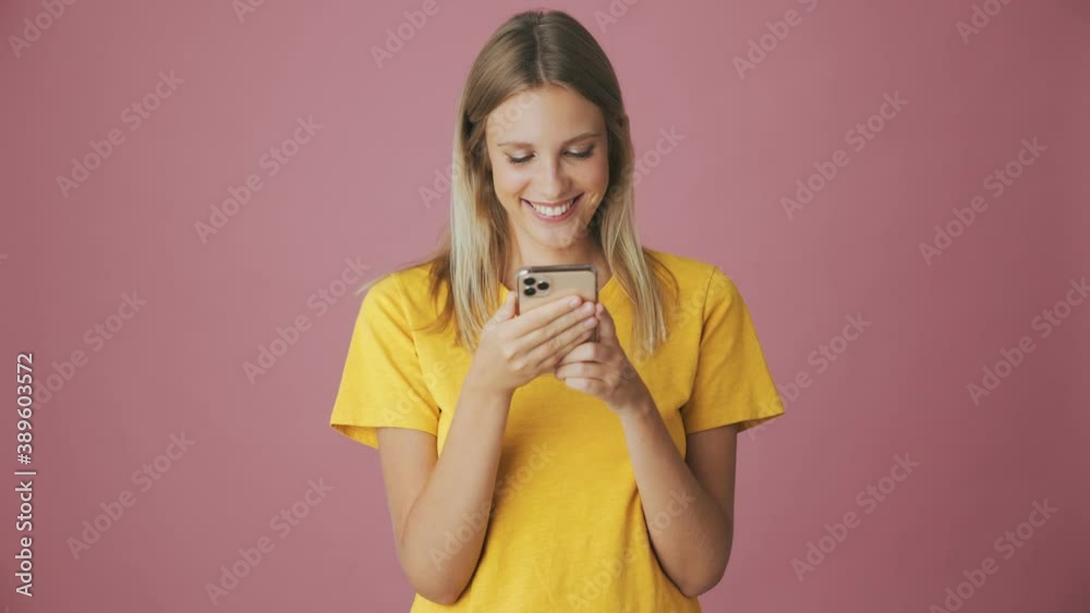 A focused blonde woman is using her smartphone standing isolated over a pink background in the studio A focused blonde woman is using her smartphone standing isolated over a pink background in the studio