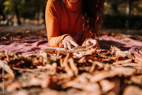 Close up. Selective focus. A woman reading a book in a park in a autumnal day. She has a maroon nails and she is wearing an orange sweater. Lifestyle autumnal concept