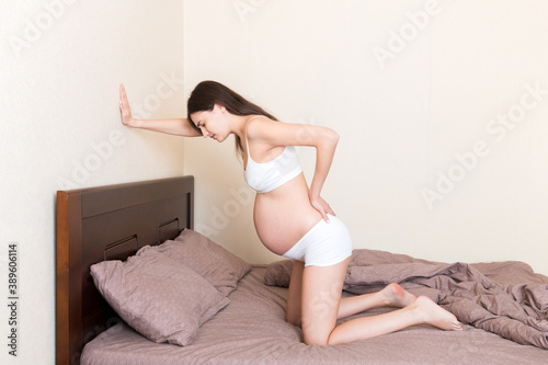 beautiful pregnant woman touching her tummy and keeping one hand on her back at home on bed