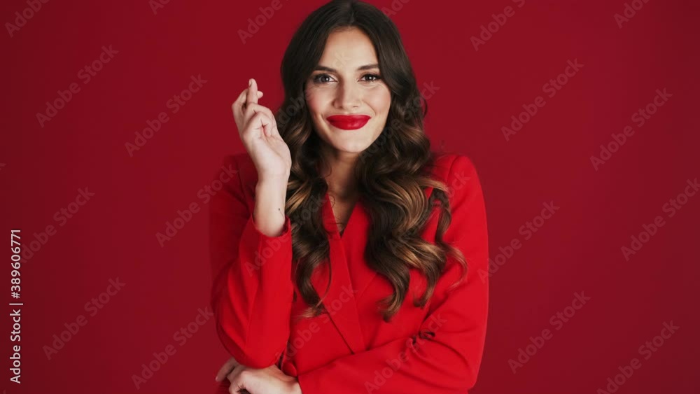 A smiling woman in a red blazer is crossing her fingers while making a wish standing isolated over red background in the studio A smiling woman in a red blazer is crossing her fingers while making a wish standing isolated over red background in the studio