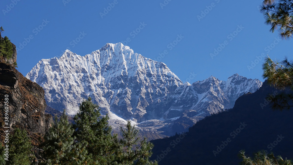 Majestic mountain Thamserku (6,623 m) in the Himalayas seen from Mount Everest Base Camp Trek near Manjo, Nepal framed by coniferous trees on a sunny day with clear sky.