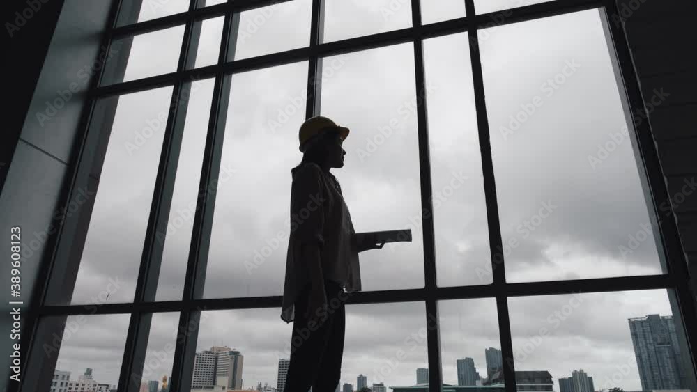 Rear view of silhouette engineer and architects woman wear security helmet with blueprint paper standing in construction site looking through full-length window at cityscape with skyscrapers.