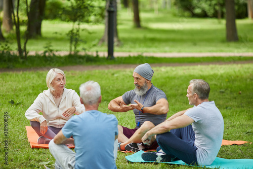 Group of active senior people gathered together in city park sitting on mats listening to their yoga trainer