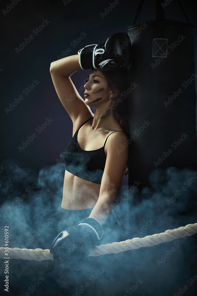 sporty woman in black boxing gloves leaning on punching bag in dark background. woman resting after workout