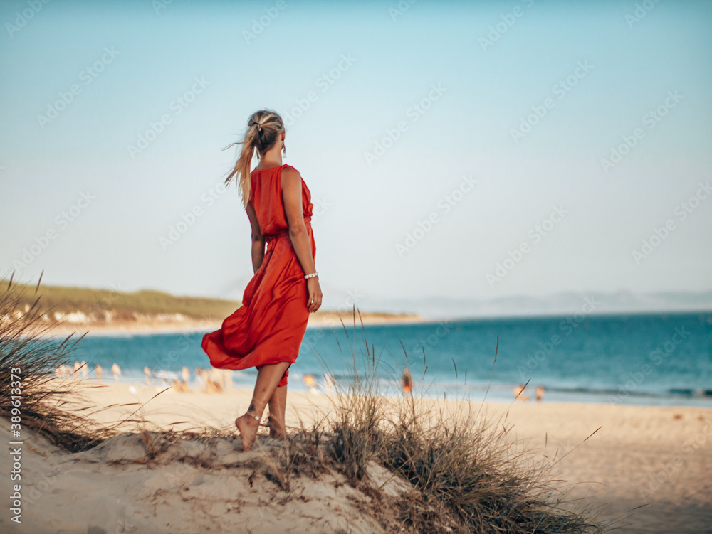 Pretty Blond Girl wearing a long red dress on the beach, playing with sand and looking towards the ocean