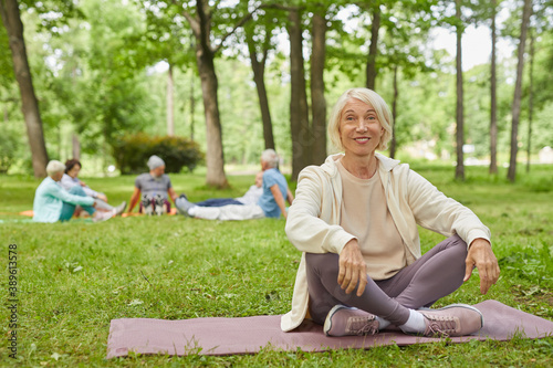 Full body shot of happy senior woman with gray hair sitting on mat in park with legs crossed smiling at camera