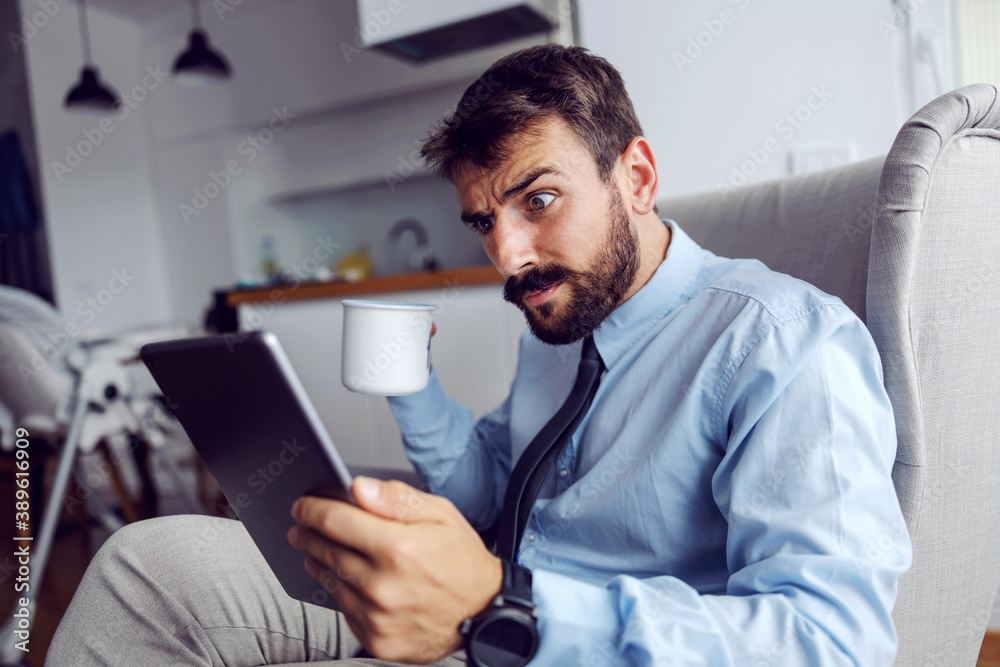Young shocked father in suit sitting at home in chair, drinking coffee and reading mails. Young shocked father in suit sitting at home in chair, drinking coffee and reading mails.