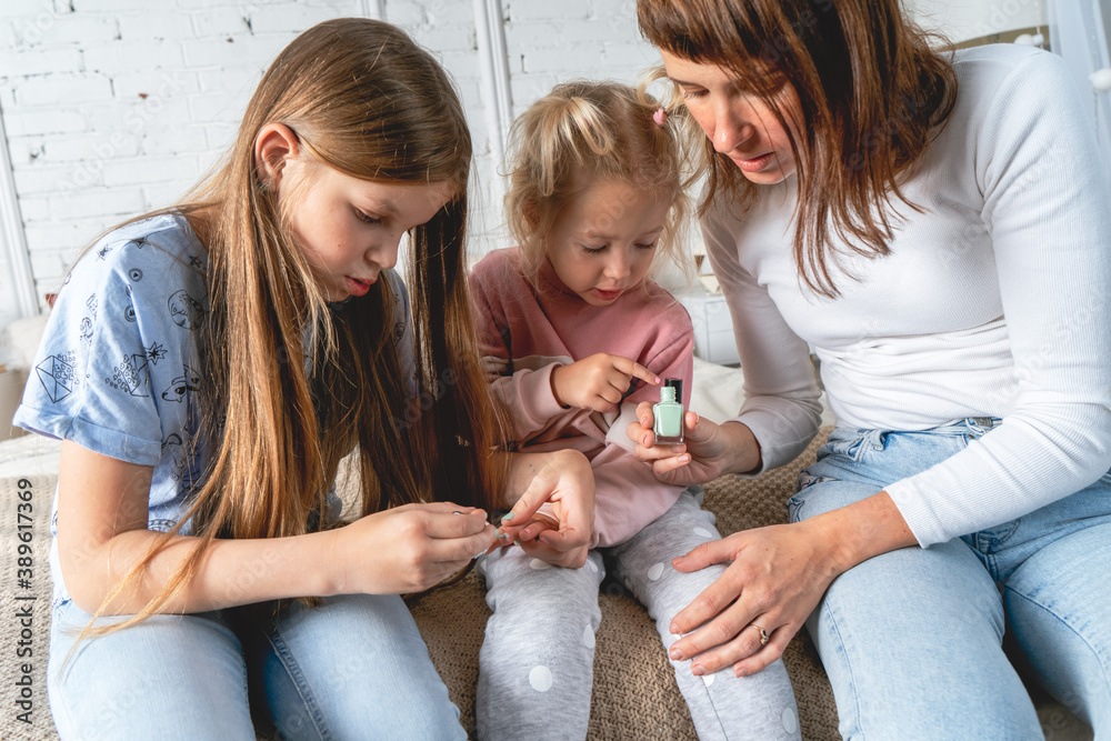 Mom and daughters sit on the bed in the bedroom and paint each other's nails with nail Polish.