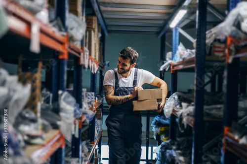 Hardworking smiling tattooed bearded worker relocating boxes while walking in storage of import and export firm.