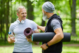 Two sporty mature adult men standing somewhere in park holding mats talking about something