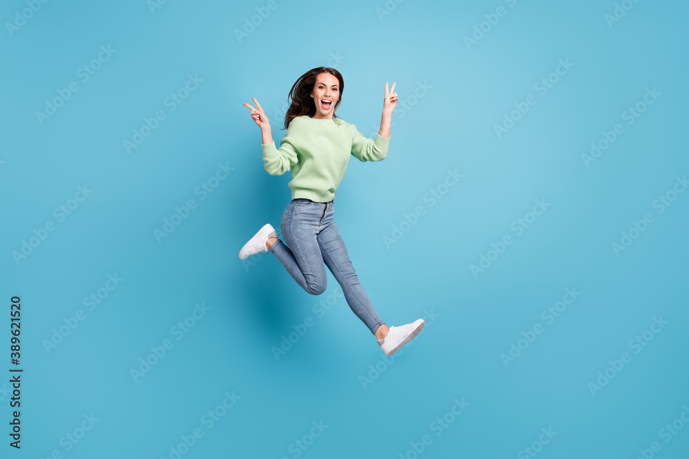 Full length body size side profile photo of pretty young female student demonstrating v-sign gesture with both hands jumping up smiling isolated on vibrant blue color background