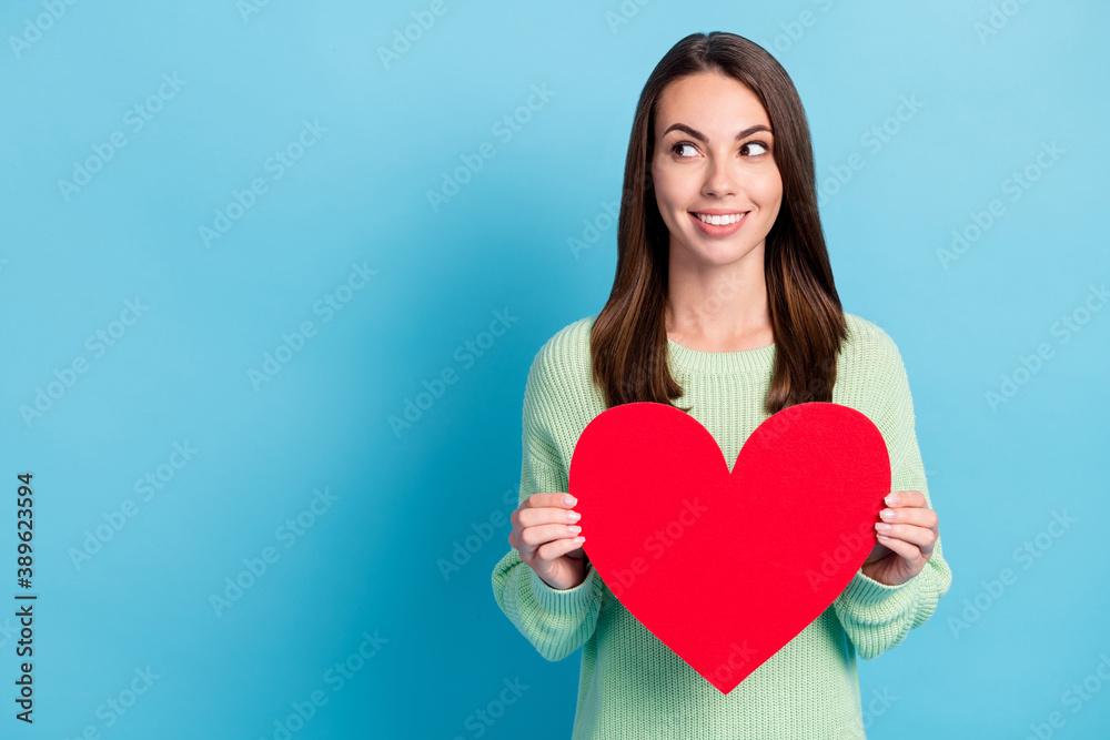 Photo portrait of curious pretty girl holding red symbol of love big heart wearing green jumper smiling looking up isolated on vibrant blue color background with empty space Photo portrait of curious pretty girl holding red symbol of love big heart wearing green jumper smiling looking up isolated on vibrant blue color background with empty space