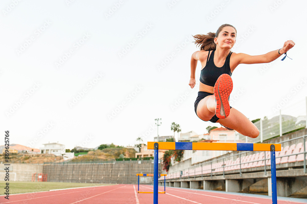 Fit female teenager athlete hurdler running jumping over hurdles - Copy space