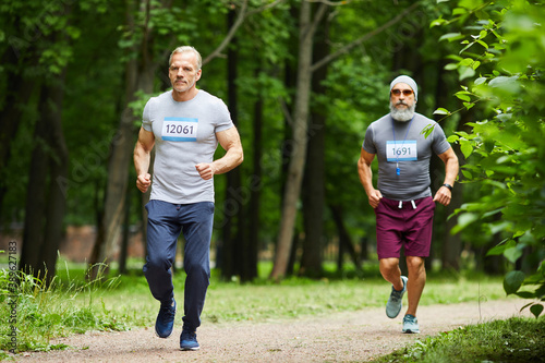 Fototapeta Naklejka Na Ścianę i Meble -  Two handsome sporty aged men taking part in marathon race in city park on summer day, wide shot