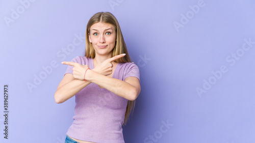 Young blonde woman isolated on purple background points sideways  is trying to choose between two options.