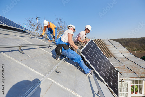 Male workers installing stand-alone solar photovoltaic panel system. Electricians lifting blue solar module on roof of modern house. Alternative energy ecological concept.