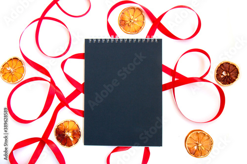 Bright festive red ribbon and curl and a notebook with black pages and slices of dried lemon isolated on white background. List of goals for the new year. Blank template