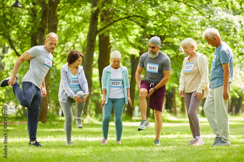 Group of senior men and women taking part in marathon training stretching legs just before running in park Group of senior men and women taking part in marathon training stretching legs just before running in park