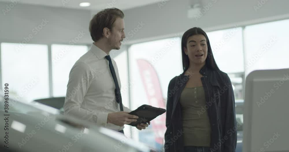 Car sales person talking to customer in car dealership showroom, looking at digital tablet