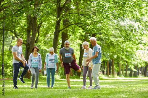 Long shot of active seniors taking part in summer marathon race standing together somewhere in park doing stretching exercise