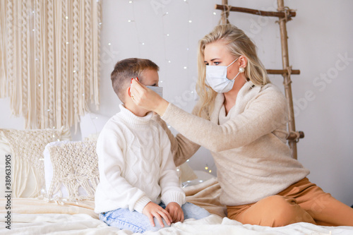 A beautiful mother and son in medical masks are having fun at home near a Christmas tree in a white interior. Family happiness, holiday, joy, vacation, games with a woman. New Year's preparations.