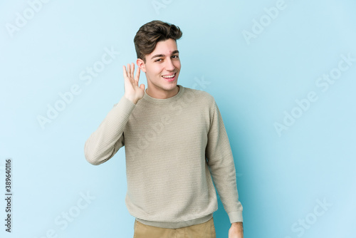 Young caucasian man isolated on blue background trying to listening a gossip.