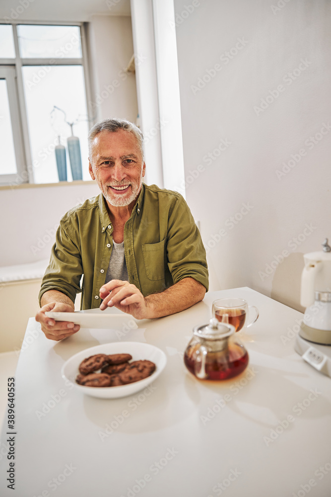 Joyous elderly man with a tablet having tea with biscuits Joyous elderly man with a tablet having tea with biscuits