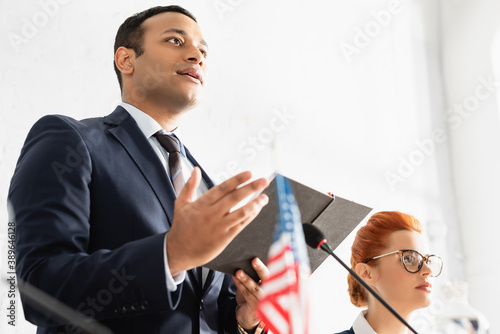 Low angle view of indian politician pointing with hand and holding notebook, standing near female colleague on blurred foreground