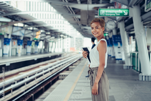 Woman standing at train station, holding digital tablet and looking and smiling at camera