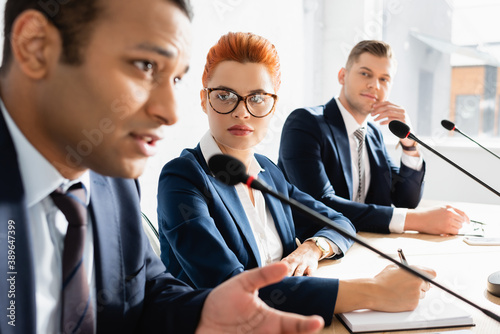 Redhead female politician looking at indian colleague speaking in microphone during political party meeting in boardroom on blurred foreground photo