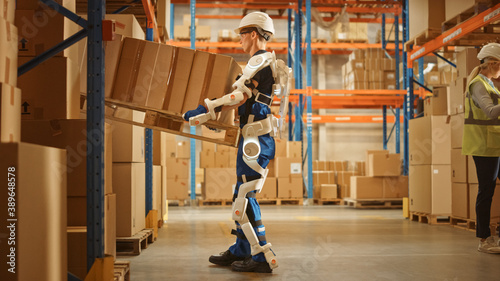 High-Tech Futuristic Warehouse: Worker Wearing Advanced Full Body Powered exoskeleton, Lifts Heavy Pallet full of Cardboard Boxes. Delivery Exosuit amplifies strength. photo