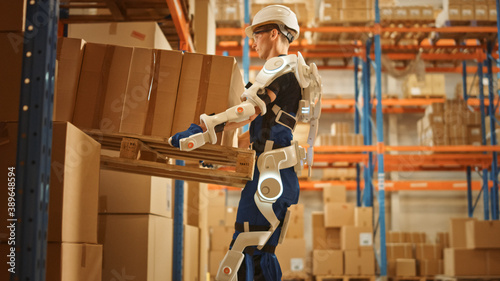 High-Tech Futuristic Warehouse: Worker Wearing Advanced Full Body Powered exoskeleton, Lifts Heavy Pallet full of Cardboard Boxes. Delivery Exosuit amplifies strength. photo