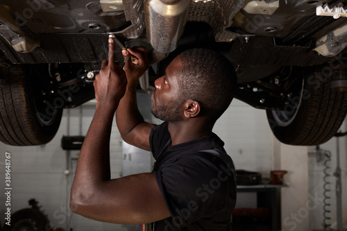 young male car mechanic in uniform checking car in automobile service with lifted vehicle, handsome hardworking guy working under car condition on lifter. automotive car repair concept