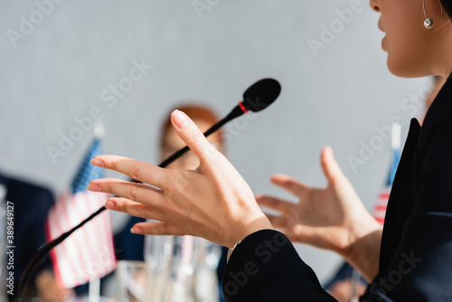 Cropped view of female politician gesturing  while speaking in microphone with blurred woman on background