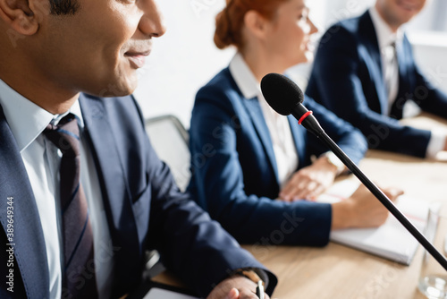 Positive indian politician sitting near microphone with blurred colleagues on background photo