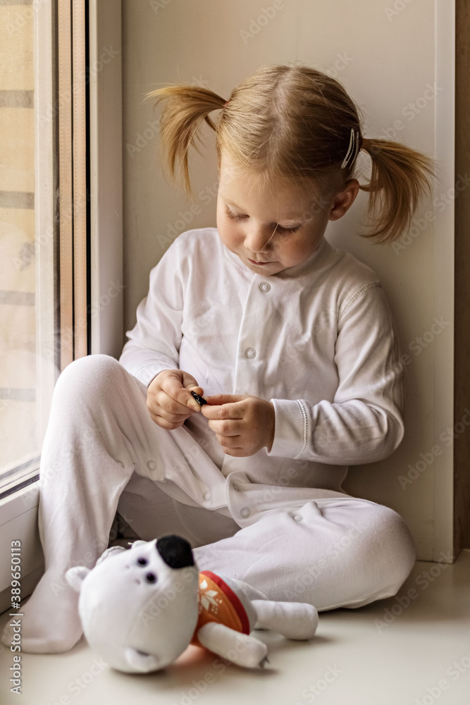 A little girl in white clothes plays with soft toys by the window. Self-isolation and quarantine content during the coronavirus pandemic in the world. A little girl in white clothes plays with soft toys by the window. Self-isolation and quarantine content during the coronavirus pandemic in the world.