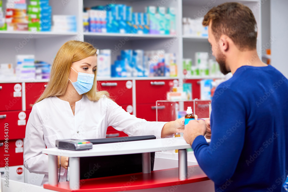 young professional druggist give medications to customer in modern drugstore, consult and recommend, wearing medical mask protecting from coronavirus covid-19